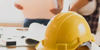 Construction helmet on table with two people reviewing plans in the background